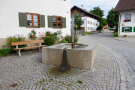 Der Dorfbrunnen in Stötten in der Ortsmitte mit einer Sitzbank.