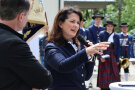 Eine Frau mit blauem Jacket hält ein Mikrofon in der Hand und spricht zu den Gästen. Im Hintergrund stehen Musiker der Blaskapelle.