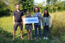 Zwei Frauen und zwei Männer stehen auf einer Wiese und halten ein Schild mit der Aufschrift „FlurNatur“.