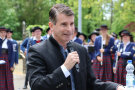 Ein Mann mit dunklem Trachtenjanker hält ein Mikrofon in der Hand und spricht zu den Gästen. Im Hintergrund stehen Musiker der Blaskapelle.