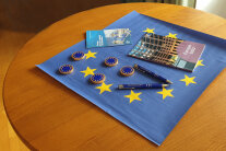 Ein Stehtisch ist mit einer Euro-Tischdecke, Eurotalern, Eurokugelschreibern sowie mit zwei Informationsheften über Europa geschmückt.