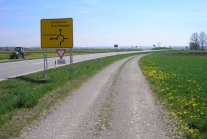 Ein landwirtschaftlicher Kiesweg neben einer asphaltierten Straße. Auf dem Grünstreifen dazwischen steht ein gelber Verkehrswegweiser. 