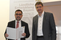 Staatsminister Klaus Holetschek hält eine Broschüre, neben ihm steht Amtsleiter Christian Kreye.