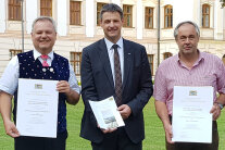 Drei Männer stehen nebeneinander und halten eine Urkunde in der Hand.