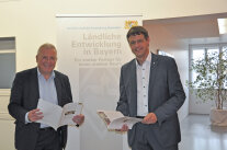 Zwei Männer halten eine Broschüre, im Hintergrund steht ein Rollup mit der Aufschrift Ländliche Entwicklung in Bayern. 