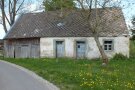 Ein altes einstöckige Gebäude mit undichtem Dach steht hinter einem Baum ohne Blätter.