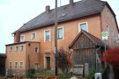 Rückwärtige Ansicht des Schul- und Rathauses vor der Sanierung mit roter Fassade und weißen Fensterumrahmung mit Buswartehäuschen aus Holz im Vordergrund