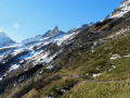 Ansteigender Alpweg, der etwa hangparallel verläuft. Im Hintergrund noch etwas verschneite Berggipfel.