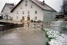 Dirlewang bei Hochwasser: Zwei Wohnhäuser umgeben von Wasser. Haus im Vordergrund verhindert mit Sandsäcken Wassereintritt durch die beiden Eingangstüren. Überschwemmt sind auch Wege und Mistlege.