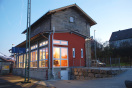 Dreigeschossiges Bahnhofsgebäude - ein Sandsteinbau - mit Satteldach. Auf den Fassaden steht groß Hagenbüchach. Im Vordergrund ein eingeschossiger Anbau mit dunkelroter Fassade und langen Fensterfronten.