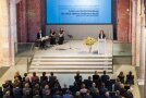 Das Publikum schaut gespannt auf die Bühne mit Kirchenfassade. Auf der Bühne steht die Ministerin mit der Band.