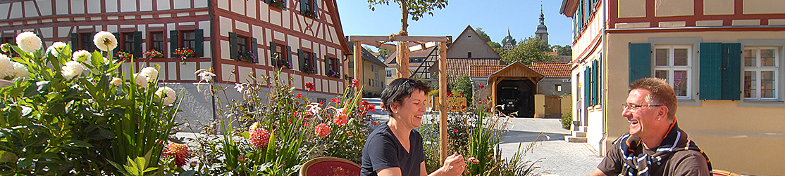 Zwei Personen unterhalten sich und sitzen dabei am Dorfplatz mit Fachwerkgebäuden im Hintergrund